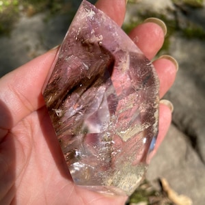 May include: A faceted, translucent crystal with shades of purple, brown, and clear. The crystal is held in a hand, showcasing its geometric shape and natural inclusions. The crystal's surface reflects light, highlighting its polished facets.