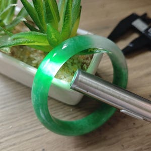 May include: A smooth, circular bangle bracelet crafted from translucent green and white jadeite stone. The bangle features a polished surface with natural color variations, illuminated by a silver-toned metal flashlight held against the side.