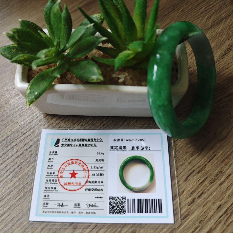 May include: A smooth, polished green jade bangle bracelet with translucent and opaque color variations. The bracelet is accompanied by a white authentication certificate featuring a red circular seal, a barcode, and technical data including 55.0g.