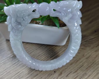 May include: A light green jade bracelet with two dragon heads facing each other. The bracelet is carved with scales and details. The background includes a white pot with green plants.