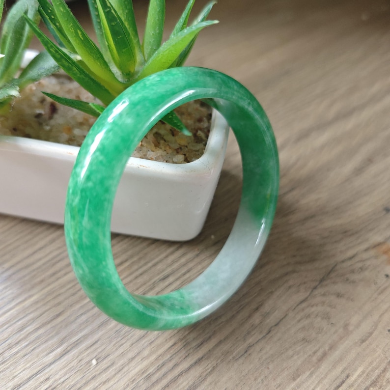 May include: A smooth, circular bangle bracelet crafted from polished green jade stone. The bangle features a vibrant, mottled emerald green coloration that transitions into a lighter, translucent white section along one side of the rounded band.