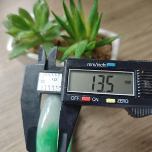 May include: A smooth, translucent green and white jade bangle bracelet being measured by a black digital caliper. The digital display clearly shows a measurement of 13.5 mm, indicating the thickness of the rounded stone band.