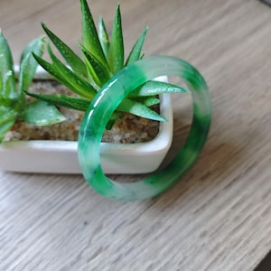 May include: A smooth, polished bangle bracelet featuring a translucent green and white marbled pattern. The circular accessory has a glossy finish and displays varying shades of emerald and light green throughout its solid, continuous band structure.