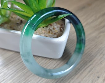 May include: A jade bangle bracelet with a gradient of colors, transitioning from a light, almost translucent green to a deep, dark green. The smooth, polished surface reflects light, highlighting the natural variations in the stone. The bracelet is circular.