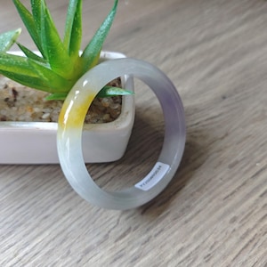 May include: A smooth, circular jade bangle bracelet featuring a translucent light grey base with a distinct patch of vibrant yellow and a subtle hint of lavender. A small white rectangular sticker with black text is attached to the inner band.