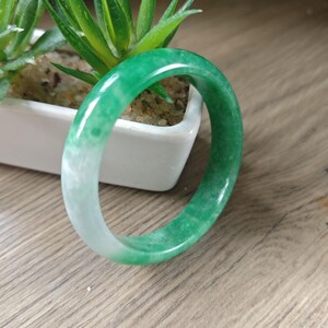 May include: A smooth, polished bangle bracelet featuring a gradient of vibrant green and translucent white tones. The circular jewelry piece has a rounded profile and a glossy finish, showcasing natural color variations throughout the material.