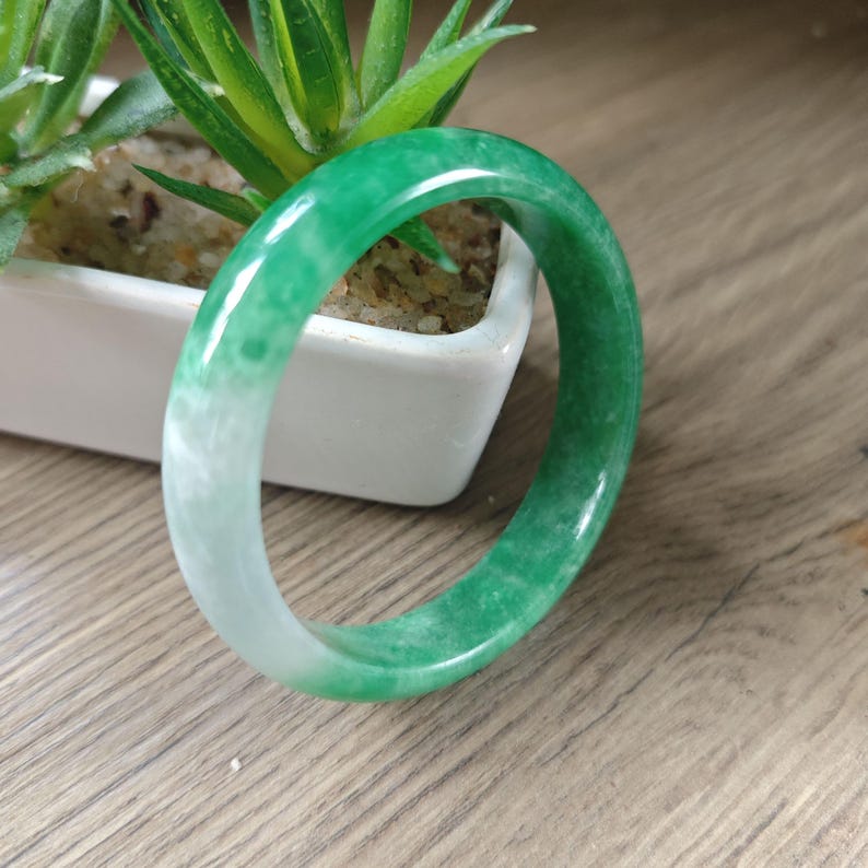 May include: A smooth, polished bangle bracelet featuring a gradient of vibrant green and translucent white tones. The circular jewelry piece has a rounded profile and a glossy finish, showcasing natural color variations throughout the material.
