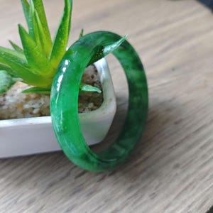 May include: A smooth, polished bangle bracelet featuring a vibrant, translucent emerald green colour with darker green mottled patterns throughout the circular band, showcasing a glossy finish that reflects light across its rounded surface.