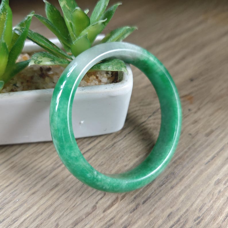 May include: A smooth, polished jade bangle bracelet featuring a vibrant green color with subtle white marbling throughout. The circular accessory has a rounded profile and a glossy, reflective surface that catches the light, showcasing the natural stone texture.