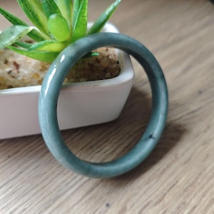 May include: A smooth, jade green bangle bracelet. The polished surface reflects light, showcasing the natural variations in color. The bracelet is a solid, circular band, perfect for everyday wear. The background includes a small white planter with a green plant.