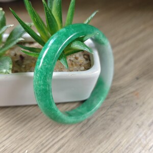 May include: A smooth, polished, circular bangle bracelet featuring a vibrant green color with subtle white mottled patterns throughout the stone, showcasing a translucent quality and a rounded, continuous band design.