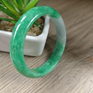 May include: A smooth, polished bangle bracelet crafted from green and white mottled stone. The circular band features a glossy finish with varying shades of vibrant green and translucent white patches distributed throughout the solid material.