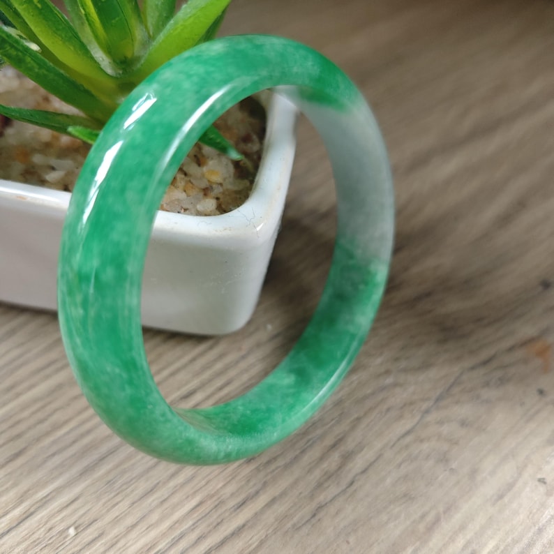 May include: A smooth, polished bangle bracelet crafted from green and white mottled stone. The circular band features a glossy finish with varying shades of vibrant green and translucent white patches distributed throughout the solid material.