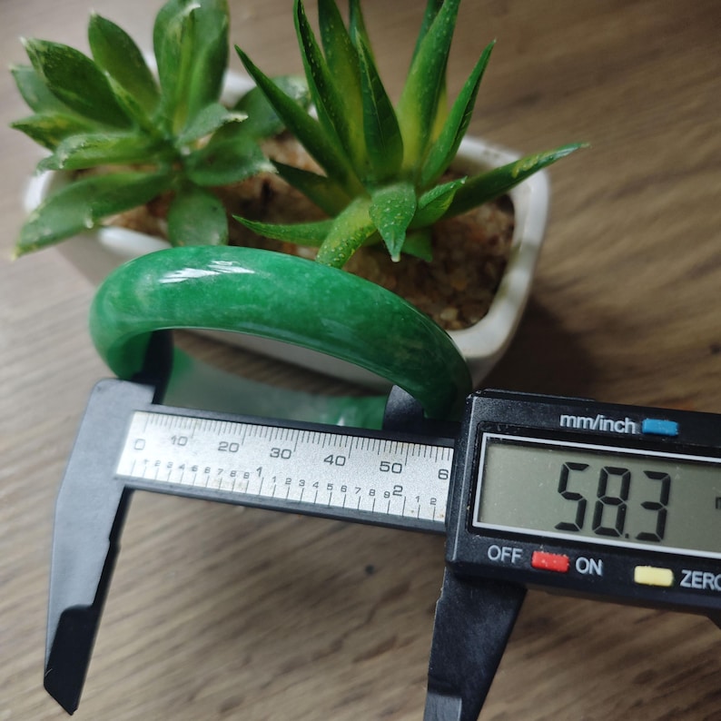 May include: A smooth, solid green jade bangle bracelet being measured by a black digital caliper. The digital display clearly shows a measurement of 58.3 mm. The bangle has a polished, glossy finish and a consistent, rounded cylindrical shape.