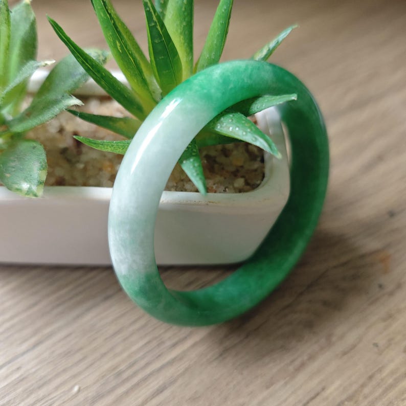 May include: A smooth, circular bangle bracelet crafted from polished stone, featuring a gradient of vibrant emerald green transitioning into a translucent pale white. The bangle has a rounded profile and a glossy, reflective surface finish.