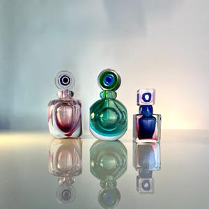 May include: Three colorful glass perfume bottles with unique stopper designs. The bottles feature shades of pink, green, and blue, with decorative accents. The bottles are reflected on a glossy surface.