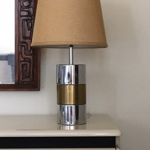 May include: A chrome and brass table lamp with a beige lampshade. The lamp has a cylindrical base with a stacked design.