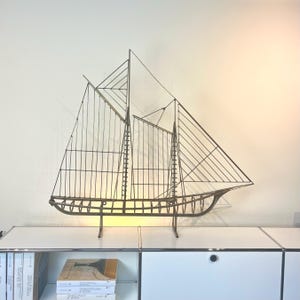 May include: A decorative metal sailboat sculpture. The sculpture is made of thin, dark metal rods, forming the hull, masts, and sails of the ship. The sculpture is displayed on a white cabinet against a cream-colored wall.
