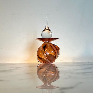 May include: A hand-blown glass bottle with a swirl design in a light amber color. The bottle has a clear glass stopper in the shape of a teardrop.