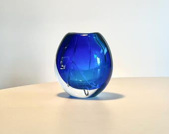 May include: A round, blue glass vase with a clear base. The vase has a modern design with a smooth, glossy finish. The blue color is vibrant and eye-catching, making it a decorative piece for any home.
