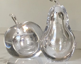May include: Two clear glass sculptures of fruit, an apple and a pear. The sculptures are made of clear glass and have a smooth, polished finish.