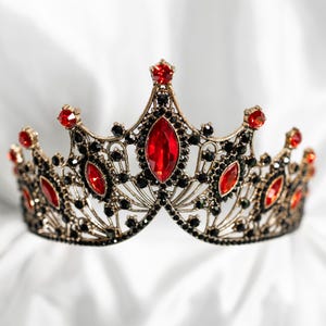 Peut inclure: Une couronne ornée avec une monture en métal doré, ornée de pierres précieuses rouges et noires. La couronne présente une grande pierre rouge centrale et de petits accents rouges, avec des pierres noires remplissant les espaces. Le design est complexe et détaillé.