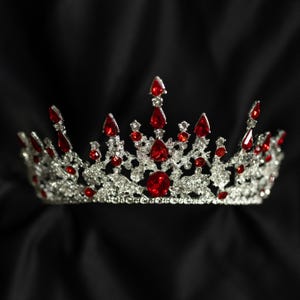May include: A silver-colored crown with numerous red gemstones. The crown features a symmetrical design with various sizes and shapes of red jewels, including teardrop and round cuts. The background is a solid black.