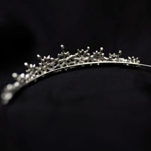 May include: A silver tiara with a delicate, intricate design. The tiara is made of metal and has a row of small, sparkling stones.