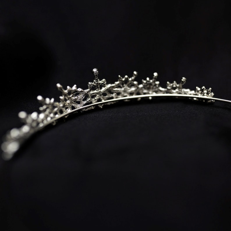 May include: A silver tiara with a delicate, intricate design. The tiara is made of metal and has a row of small, sparkling stones.