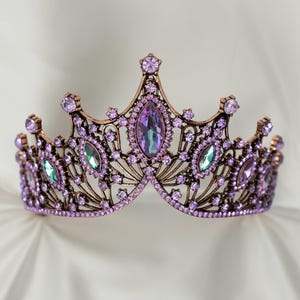 Peut inclure: Une couronne décorative avec une monture en métal de couleur bronze. La couronne est ornée de nombreuses pierres précieuses violettes et vertes irisées de différentes tailles. La pierre centrale est de forme ovale.