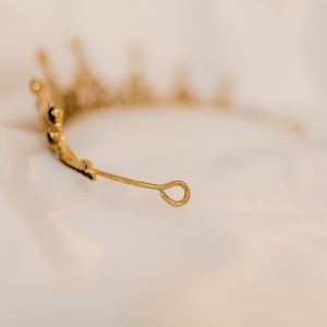 May include: A gold tiara with a teardrop-shaped loop at the back. The tiara is decorated with small, clear rhinestones.