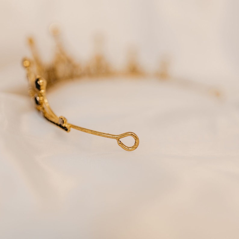 May include: A gold tiara with a teardrop-shaped loop at the back. The tiara is decorated with small, clear rhinestones.