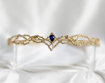 May include: A delicate gold crown with a single blue sapphire gemstone in the center. The crown is made of intricate, interwoven strands of gold.