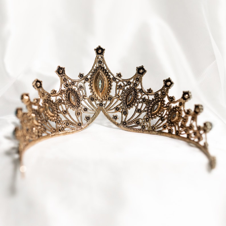 May include: A gold-toned tiara with intricate detailing and clear rhinestones. The tiara is designed with a pointed crown and delicate, flowing lines.