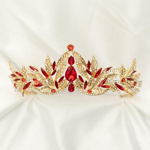 May include: A gold-toned crown with red and clear gemstones. The crown features a central teardrop-shaped red gem, surrounded by smaller red and clear stones. The design includes leaf-like accents.