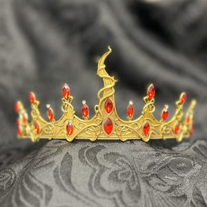 May include: A gold-colored crown with red gemstones. The crown features an ornate design with a central upward curve and is adorned with multiple red, oval-shaped gems. The crown is set against a dark, textured background.
