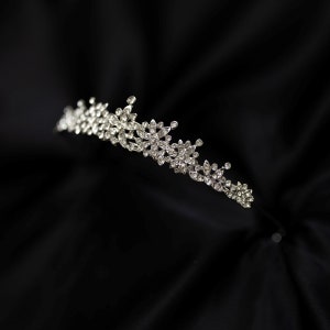 May include: A silver tiara with clear rhinestones arranged in a floral pattern. The tiara is on a black fabric background.