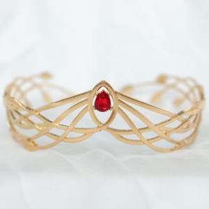 Peut inclure: Une couronne dorée avec un design complexe et entrelacé. La couronne présente une pierre précieuse rouge en forme de goutte d'eau en son centre. La fine métallurgie suggère un thème fantastique ou nuptial.