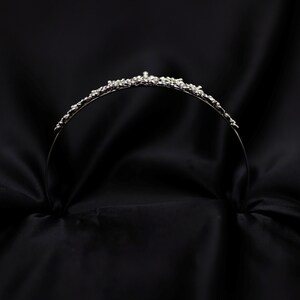 May include: A silver tiara with a delicate design of small, clear rhinestones. The tiara is on a black background.