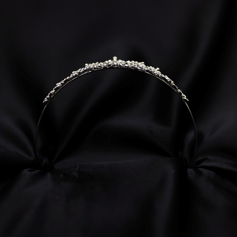 May include: A silver tiara with a delicate design of small, clear rhinestones. The tiara is on a black background.