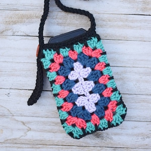 May include: A crocheted phone case with a granny square design in shades of teal, pink, blue, and purple. The case has a black strap and a brown button.