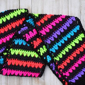 May include: A black crochet scarf with bright neon pink, green, blue, and purple stripes. The scarf is made with a simple stitch pattern and has a fringe on the ends.