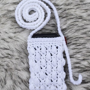 May include: White crocheted phone case with a long white strap. The case has a textured design and is perfect for carrying a smartphone.