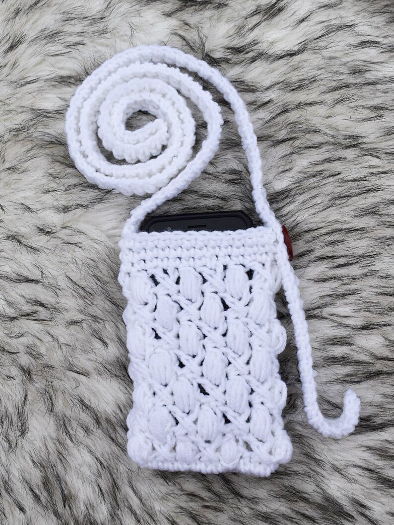 May include: White crocheted phone case with a long white strap. The case has a textured design and is perfect for carrying a smartphone.