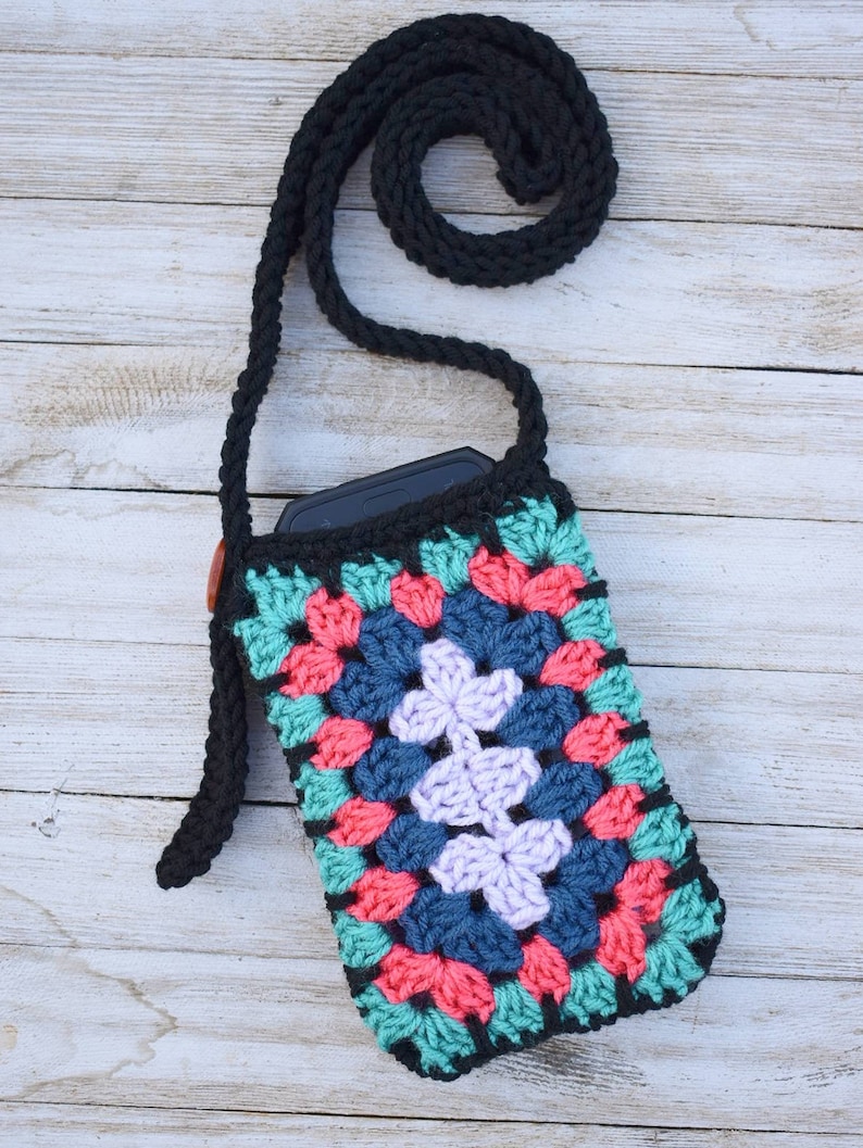 May include: A crocheted phone case with a granny square design in shades of teal, pink, blue, and purple. The case has a black strap and a brown button.
