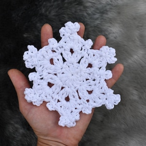 May include: A handmade white crochet snowflake. The snowflake is held in a hand, showcasing its intricate design. The snowflake is made of white yarn and has a delicate, lacy appearance. The background is a soft, textured gray.