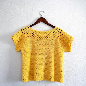 May include: A yellow crochet top with short sleeves hangs on a wooden hanger. The top features a solid crochet stitch on the lower portion and a mesh stitch on the upper portion. The top is a bright, sunny yellow colour.
