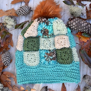 May include: A crocheted hat with a patchwork design in shades of green, blue, and beige. The hat has a fluffy brown pom-pom on top.