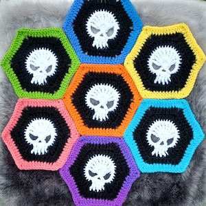 Puede incluir: Un tapiz hexagonal de ganchillo colorido con un diseño de calavera en cada sección. Las calaveras son blancas con ojos negros y están rodeadas por un contorno negro. Los hexágonos son en tonos de verde, azul, amarillo, naranja y morado.