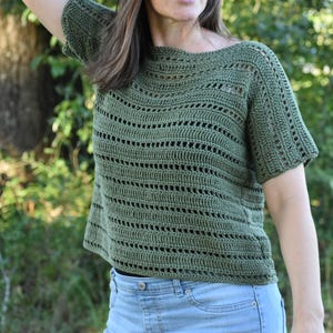 May include: An olive green crochet top with short sleeves and a boat neck. The top has a horizontal openwork design. The model is wearing light blue jeans. The background is a blurred view of trees and foliage.